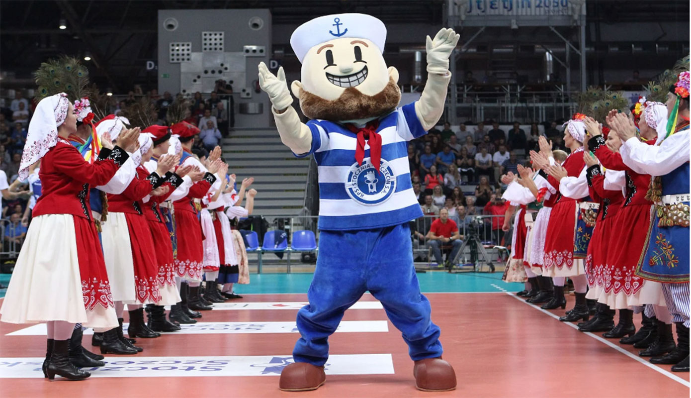 Na fotografii widnieje maskotka kostium bosmana stojący na parkiecie hali sportowej w otoczeniu tancerzy zespołu pieśni i tańca